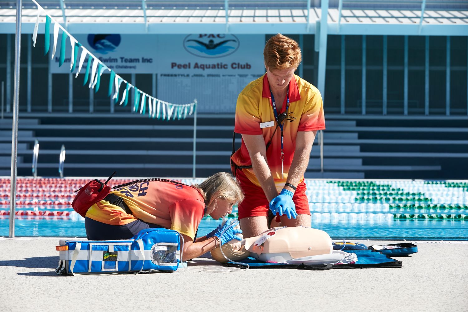Two lifesavers training CPR on a mannequin in front of a pool 